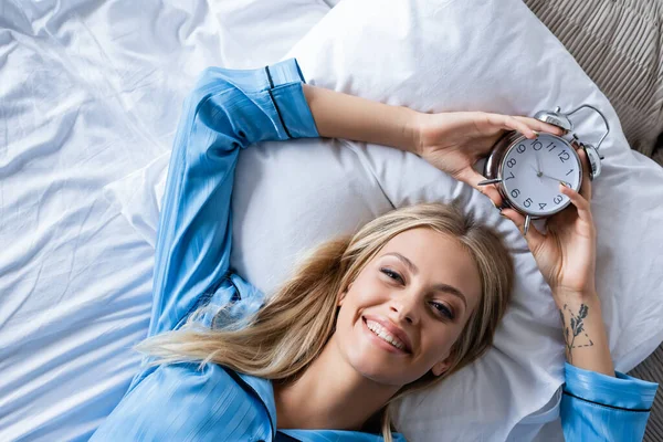 Vista superior de la mujer feliz sosteniendo reloj despertador retro y acostado en la cama - foto de stock