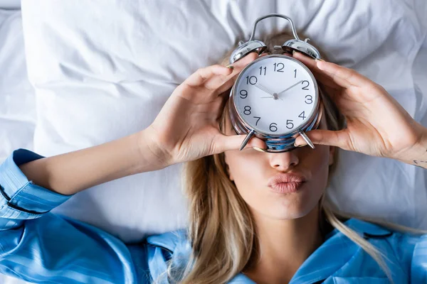 Top view of blonde woman holding retro alarm clock and pouting lips while lying on bed — Stock Photo