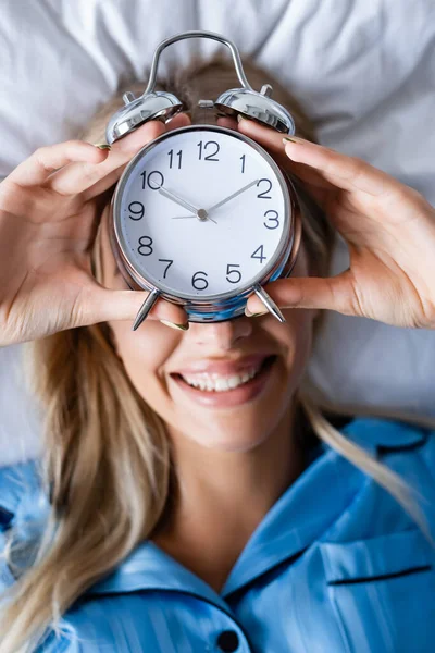 Vista superior da mulher alegre segurando despertador retro e cobrindo o rosto enquanto deitado na cama — Fotografia de Stock