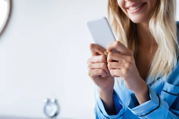 Vista ritagliata di donna allegra in pigiama che scrive su smartphone in camera da letto — Foto stock