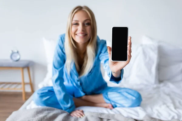 Pleased blurred woman in pajamas holding smartphone with blank screen in bedroom — Stock Photo