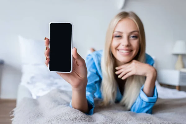 Femme floue et heureuse en pyjama tenant smartphone avec écran blanc dans la chambre — Photo de stock