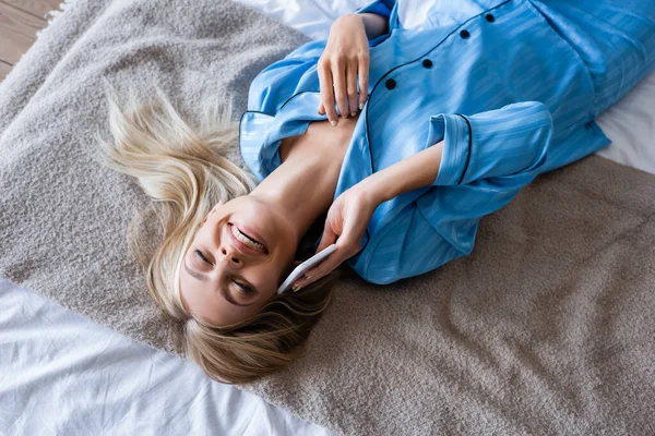 Blick von oben auf zufriedene Frau im Schlafanzug, die im Schlafzimmer auf dem Smartphone spricht — Stockfoto