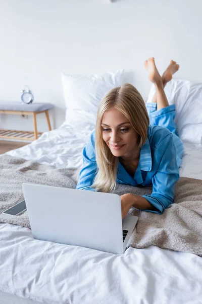 Mujer joven satisfecha en pijama usando el ordenador portátil en el dormitorio - foto de stock