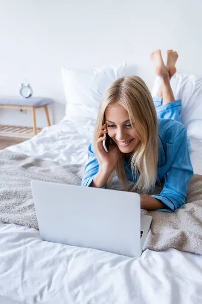 Mujer joven satisfecha en pijama usando el ordenador portátil mientras que habla el teléfono celular en dormitorio - foto de stock