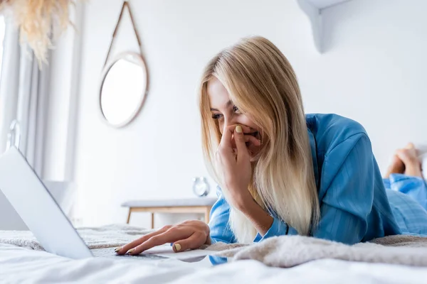 Happy blonde woman in pajamas using laptop in bedroom — Stock Photo