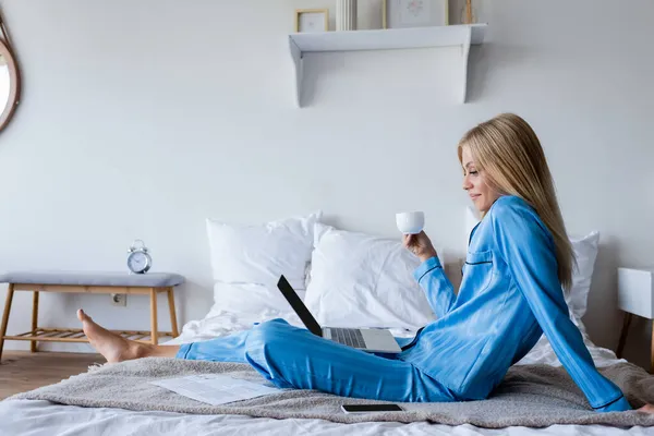 Side view of happy young woman in pajamas looking at laptop and holding cup of coffee — Stock Photo