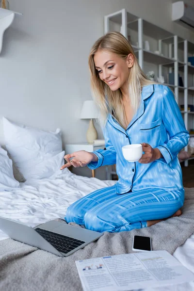 Jeune femme heureuse en pyjama tenant une tasse de café pendant l'appel vidéo — Photo de stock