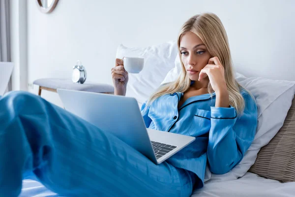 Jeune femme en pyjama utilisant un ordinateur portable et tenant une tasse de café — Photo de stock