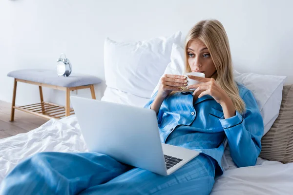 Blonde young woman in pajamas using laptop and drinking coffee — Stock Photo