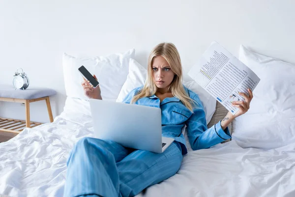 Mulher descontente segurando smartphone e jornal enquanto olha para laptop no quarto — Fotografia de Stock