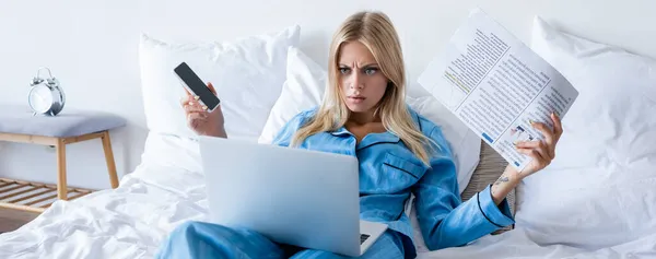 Mujer disgustada sosteniendo teléfono inteligente y periódico mientras mira el ordenador portátil en el dormitorio, pancarta - foto de stock