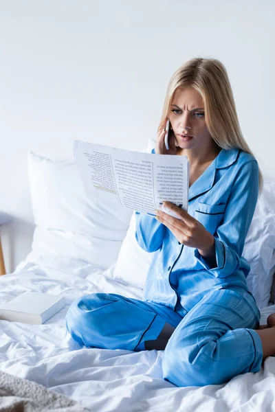 Shocked young woman in pajamas talking on smartphone and reading newspaper — Stock Photo