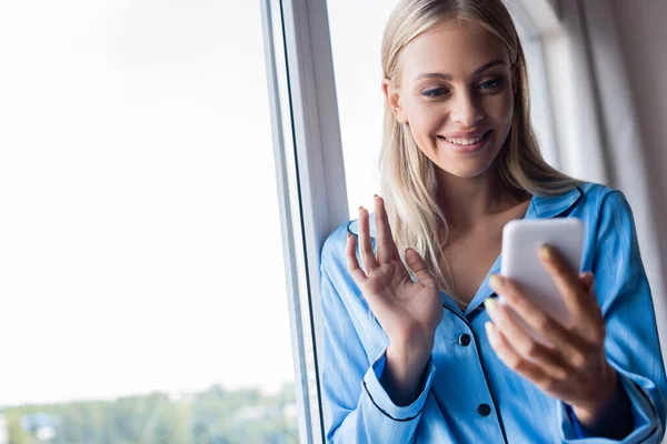 Happy young woman waving hand during video call on cellphone near window — Stock Photo