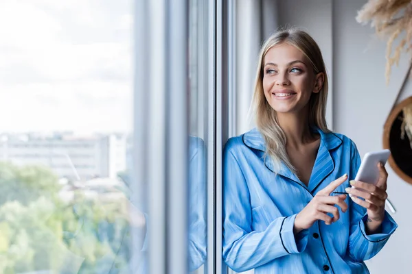 Happy young woman holding cellphone and looking at window — Stock Photo