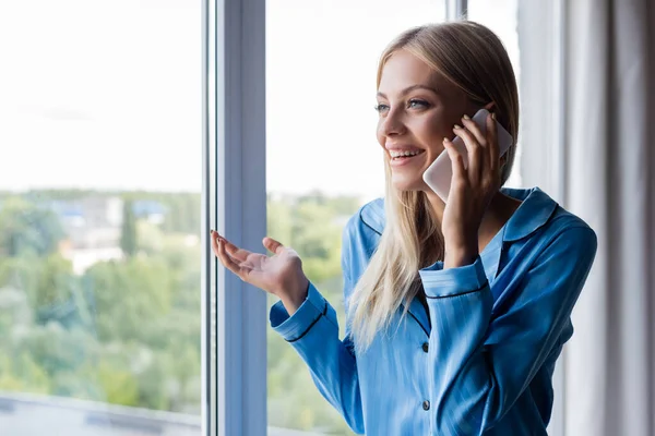 Cheerful young woman talking on cellphone near window — Stock Photo
