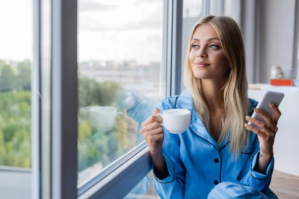 Feliz joven mujer sosteniendo el teléfono celular y la taza de café cerca de la ventana - foto de stock