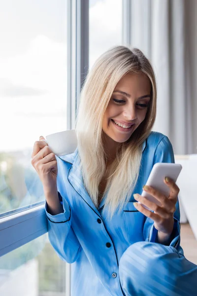 Heureuse jeune femme tenant smartphone et tasse de café près de la fenêtre — Photo de stock