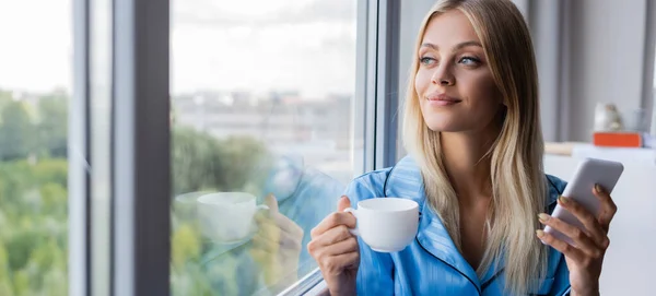 Glückliche junge Frau mit Handy und Tasse Kaffee am Fenster, Transparent — Stockfoto