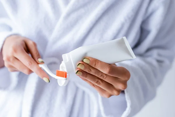 Cropped view of young woman in bathrobe holding toothbrush and toothpaste tube — Stock Photo