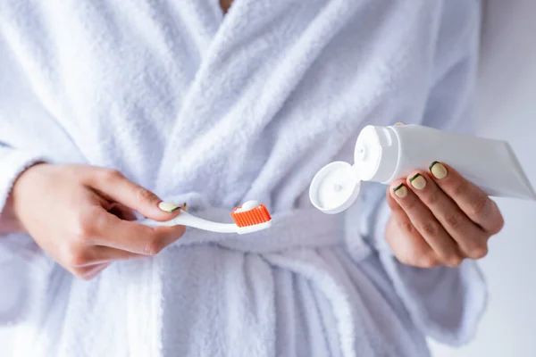 Vista parcial de la mujer en albornoz blanco sosteniendo cepillo de dientes y tubo de pasta de dientes - foto de stock