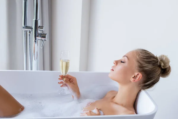 Vue latérale de la jeune femme blonde prenant un bain et tenant une coupe de champagne — Photo de stock