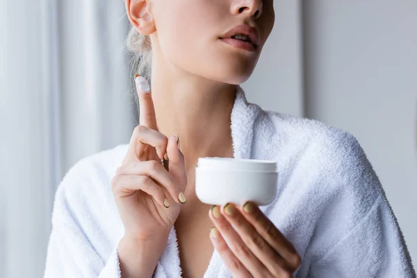 Ausgeschnittene Ansicht einer jungen Frau im Bademantel mit Behälter und Kosmetikcreme am Finger — Stockfoto