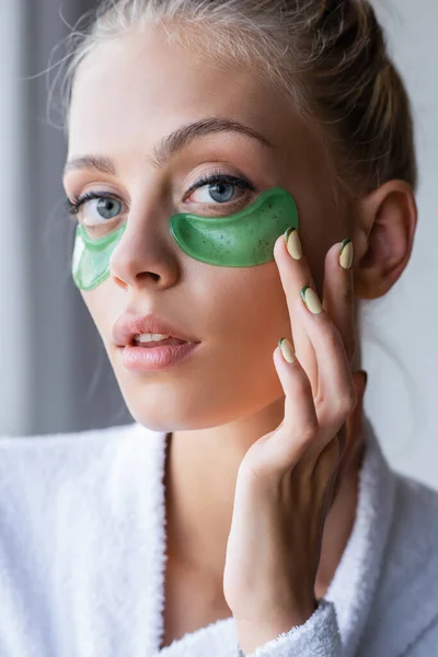 Jeune femme en peignoir appliquant des patchs d'oeil vert dans la salle de bain — Photo de stock