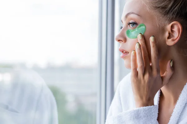 Junge Frau im Bademantel trägt grüne Augenklappe am Fenster auf — Stockfoto