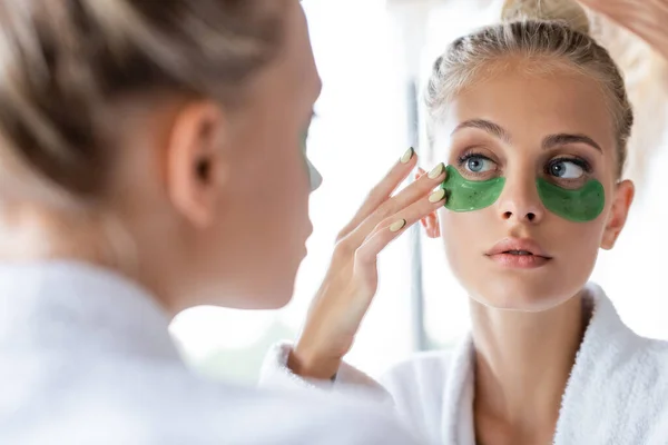 Mujer joven en albornoz aplicando parches de ojo verde cerca del espejo - foto de stock