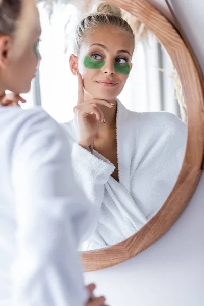 Mujer joven satisfecha en albornoz aplicando parches de ojo verde cerca del espejo - foto de stock