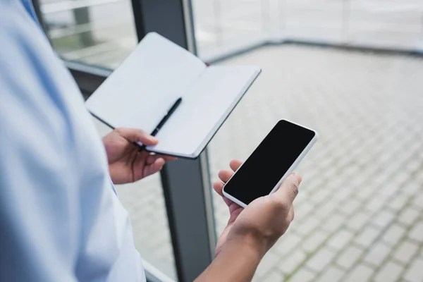 Vista recortada del hombre de negocios afroamericano sosteniendo teléfono celular con pantalla en blanco y portátil en la oficina - foto de stock