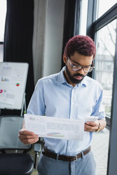 Homme d'affaires afro-américain tenant un document et utilisant un smartphone près de la fenêtre dans le bureau — Photo de stock