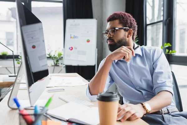 Empresário afro-americano em óculos olhando para o computador perto de café para ir e notebook no escritório — Stock Photo