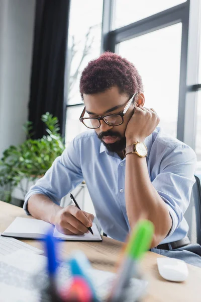 Empresário afro-americano escrevendo no notebook perto do mouse do computador no escritório — Fotografia de Stock