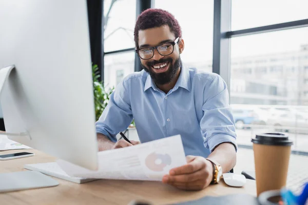 Sonriente empresario afroamericano mirando el documento cerca de la computadora y la taza de papel en la oficina - foto de stock