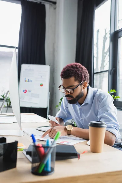 Hombre de negocios afroamericano sosteniendo teléfono inteligente y apuntando a documento cerca de la computadora en la oficina - foto de stock