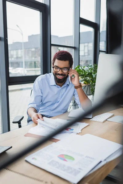 Positive african american businessman talking on smartphone and holding paper with charts in office — Stock Photo