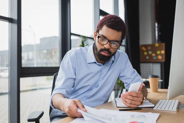 African american businessman in eyeglasses holding smartphone and documents while working in office — Stock Photo