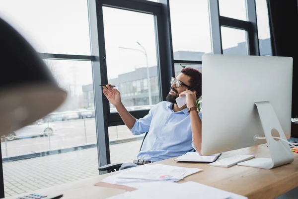 Lächelnder afrikanisch-amerikanischer Geschäftsmann mit Stift im Gespräch auf Smartphone in der Nähe von Computer und Papieren im Büro — Stockfoto