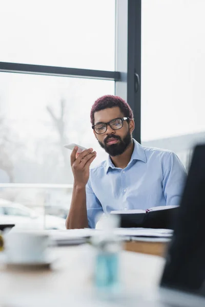 Uomo d'affari afroamericano in occhiali che tiene smartphone e guarda notebook in ufficio — Foto stock