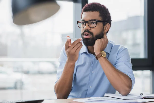 African american manager in eyeglasses recording voice message near papers in office — Stock Photo