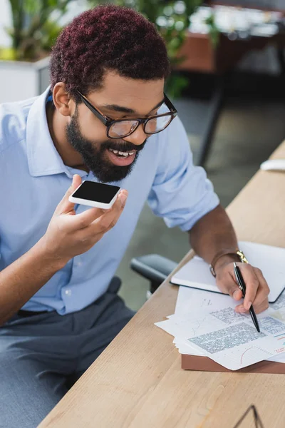 Souriant homme d'affaires afro-américain tenant smartphone et stylo près des papiers au bureau — Photo de stock