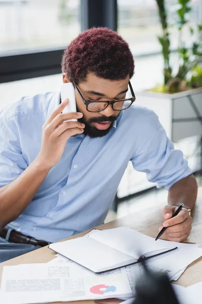 Hombre de negocios afroamericano hablando en el teléfono inteligente cerca de portátil y documentos en la oficina - foto de stock