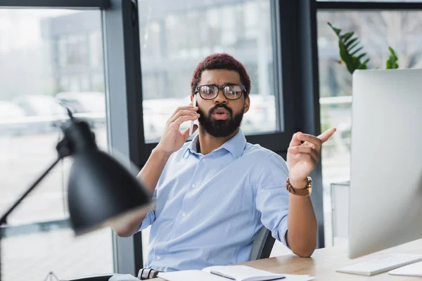 Hombre de negocios afroamericano hablando por teléfono inteligente y señalando con el dedo a la computadora en la oficina - foto de stock