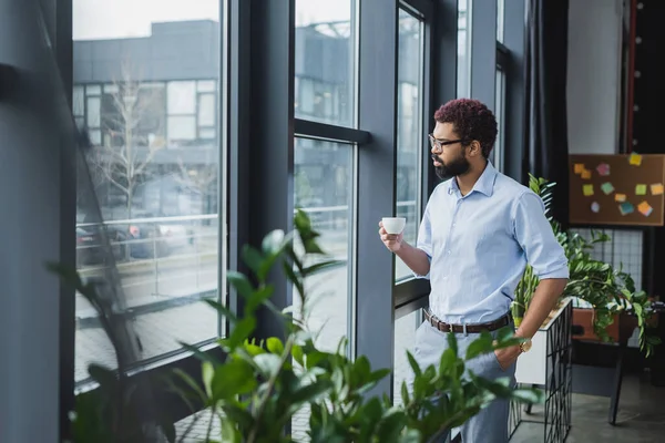 Empresario afroamericano sosteniendo taza de café cerca de la ventana en la oficina - foto de stock