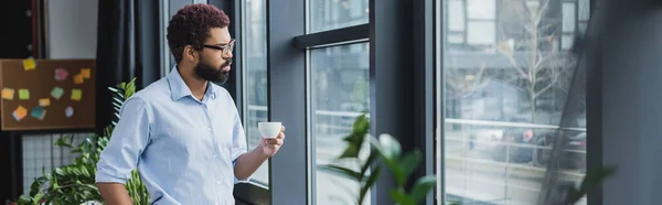 Молодой африканский американский бизнесмен в очках держа чашку возле окна в офисе, баннер — стоковое фото