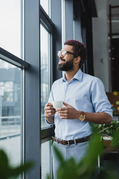 Feliz hombre de negocios afroamericano en anteojos sosteniendo taza de café cerca de la ventana en la oficina - foto de stock