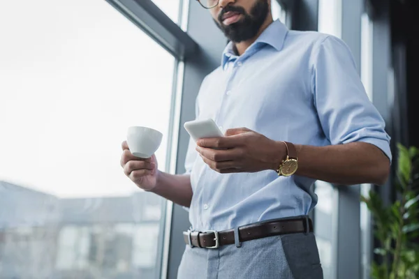 Vista de bajo ángulo del hombre de negocios afroamericano usando el teléfono celular y sosteniendo la taza de café en la oficina - foto de stock