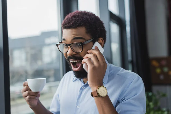 Empresário afro-americano animado falando no smartphone e segurando a xícara no escritório — Fotografia de Stock